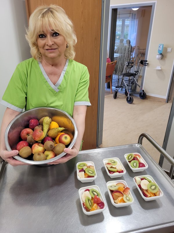 Frau hält eine Schüssel mit frischem Obst in die Kamera, daneben stehen kleine Schüsseln mit frisch zubereitetem Obstsalat.