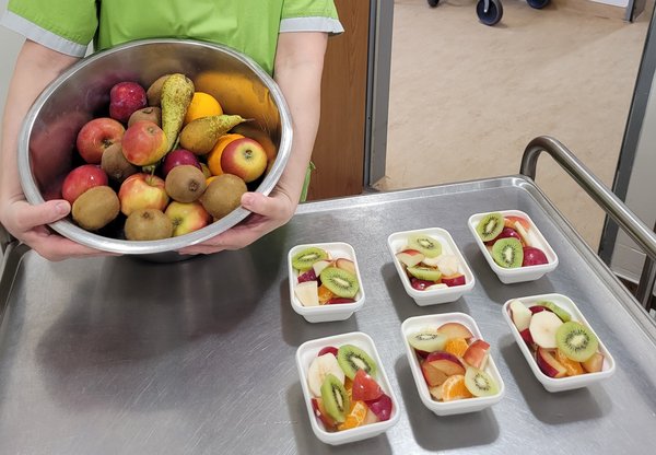 Frau hält eine Schüssel mit frischem Obst in die Kamera, daneben stehen kleine Schüsseln mit frisch zubereitetem Obstsalat.