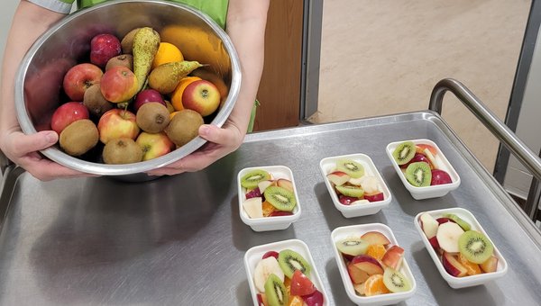 Frau hält eine Schüssel mit frischem Obst in die Kamera, daneben stehen kleine Schüsseln mit frisch zubereitetem Obstsalat.
