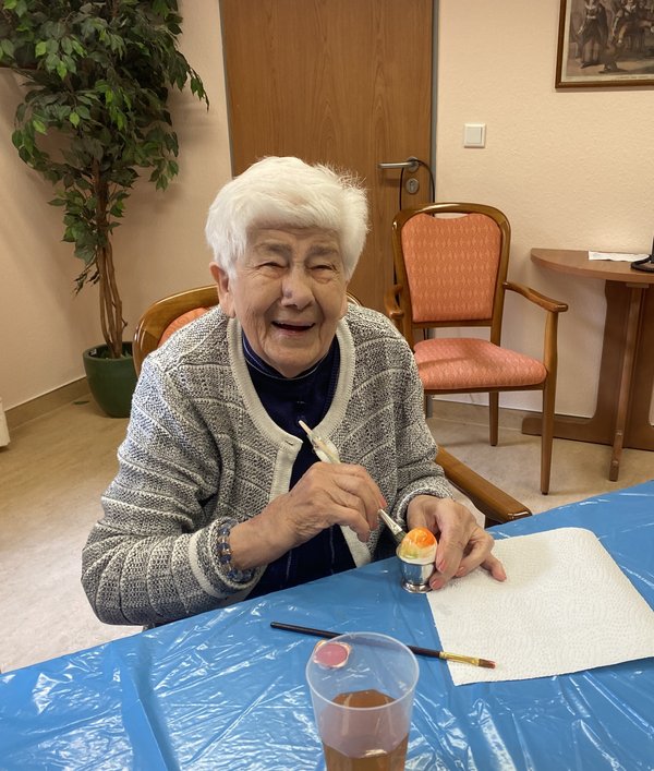 Eine Seniorin hält einen Pinsel und bemalt ein Ei. Sie lacht fröhlich in die Kamera.