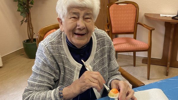 Eine Seniorin hält einen Pinsel und bemalt ein Ei. Sie lacht fröhlich in die Kamera.