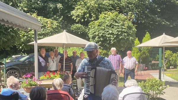 Man sieht einen Musiker mit Akkordeon, der durch das Publikum geht. Im Hintergrund sieht man einen Männerchor