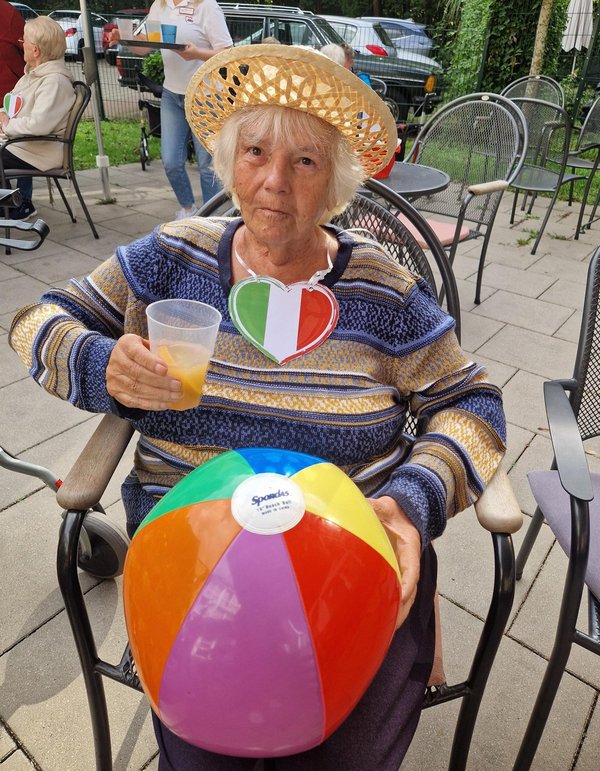 Eine Frau mit Strohhut und einem Italien-Herz-Anhänger um den Hals hält einen Wasserball in der einen und ein Getränk in der anderen Hand. Sie lacht fröhlich in die Kamera.