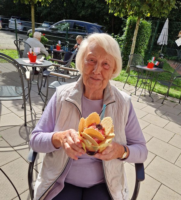 Eine Seniorin hält einen Eisbecher in der Hand und lächelt in die Kamera.