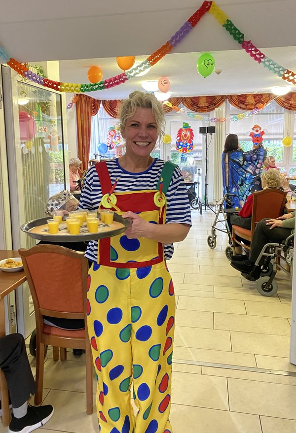 Eine Frau im Clownskostüm hält ein Tablett mit mehreren kleinen Pinnchen Eierlikör und schaut lachend in die Kamera.