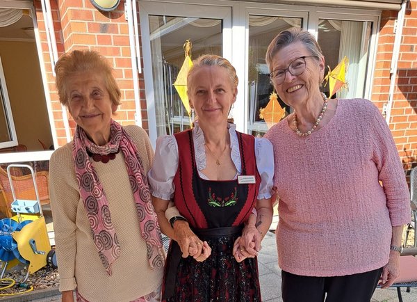 Eine Frau im Dirndl hält zwei Seniorinnen an den Händen. Alle lachen fröhlich.