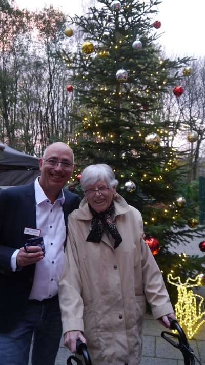 Zwei Personen stehen draußen vor einem geschmückten Tannenbaum und lächeln in die Kamera.
