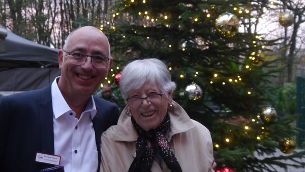 Zwei Personen stehen draußen vor einem geschmückten Tannenbaum und lächeln in die Kamera.