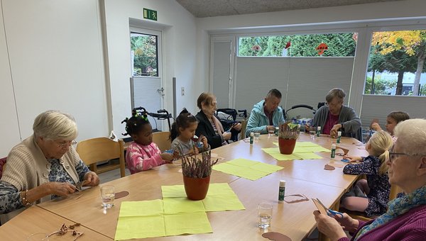 Man sieht eine Gruppe von Kindern und Seniorinnen an einem Tisch sitzen, alle schneiden konzentriert ein Stück Pappe aus.