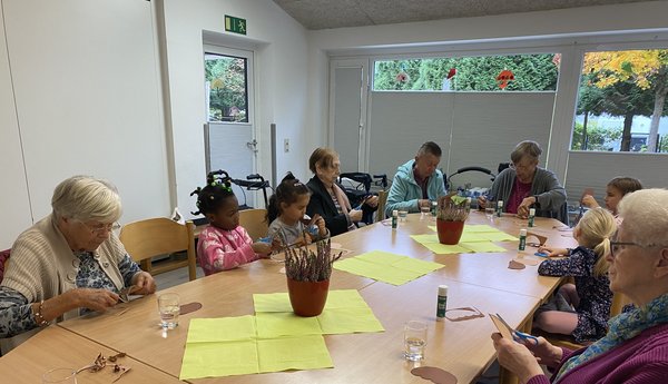 Man sieht eine Gruppe von Kindern und Seniorinnen an einem Tisch sitzen, alle schneiden konzentriert ein Stück Pappe aus.