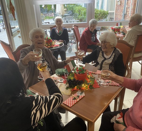 Mehrere Seniorinnen sitzen an einem italienisch gedeckten Tisch und stoßen miteinander an.