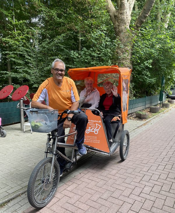 Auf dem Bild sieht man eine Fahrradrikscha, die von einem Mann gefahren wird. Hinten in der Rikscha sitzen zwei Seniorinnen, die freundlich in die Kamera winken