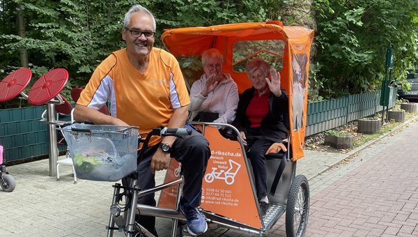 Auf dem Bild sieht man eine Fahrradrikscha, die von einem Mann gefahren wird. Hinten in der Rikscha sitzen zwei Seniorinnen, die freundlich in die Kamera winken