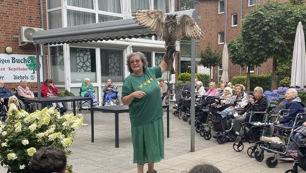 Seniorinnen sitzen im Stuhlkreis, eine Frau zeigt einen Uhu mit ausgebreiteten Flügeln. Im Vordergrund beobachten Kinder den Vogel.