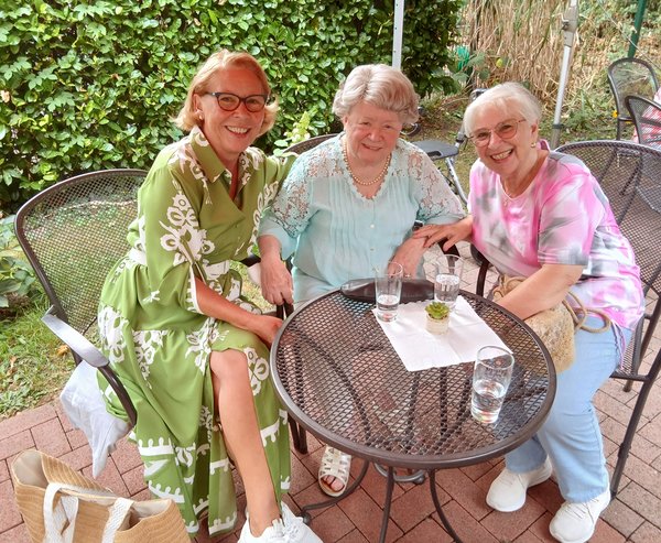 Drei Frauen sitzen gemeinsam an einem Tisch und schauen lächelnd in die Kamera.