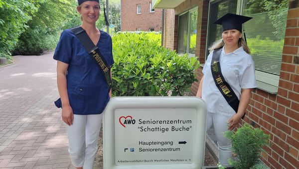 Auf dem Bild sieht man zwei Frauen, die jeweils einen Hut und eine Schärpe mit der Aufschrift ‚Prüfung bestanden‘ tragen