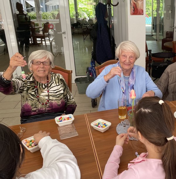 Kinder und Seniorinnen sitzen zusammen am Tisch. Die Kinder fädeln Perlen, die Seniorinnen halten lächelnd ihre fertigen Ketten in die Kamera.