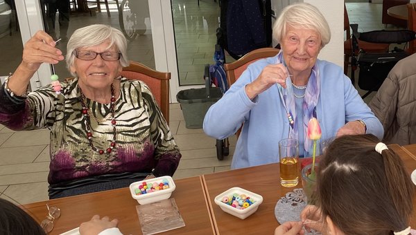 Kinder und Seniorinnen sitzen zusammen am Tisch. Die Kinder fädeln Perlen, die Seniorinnen halten lächelnd ihre fertigen Ketten in die Kamera.