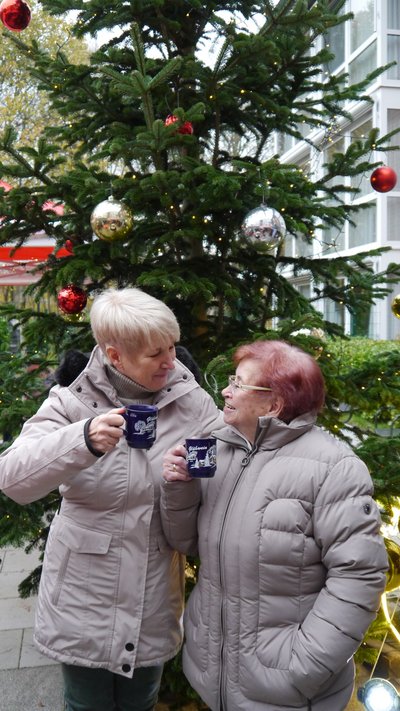Zwei Frauen stehen vor einem schön dekorierten Tannenbaum, sie halten jeweils eine Glühweintassen in der Hand und prosteten sich zu.