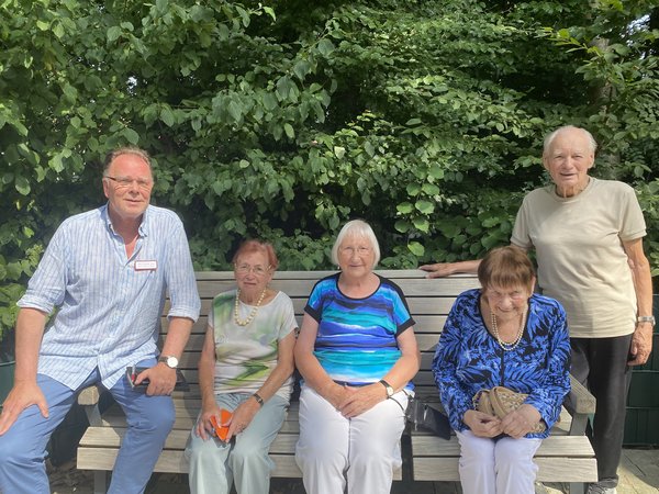 Man sieht drei Seniorinnen auf einer Bank sitzen, links und rechts steht jeweils ein Mann daneben. Alle lächeln freundlich in die Kamera.