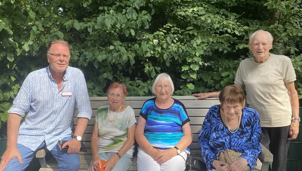 Man sieht drei Seniorinnen auf einer Bank sitzen, links und rechts steht jeweils ein Mann daneben. Alle lächeln freundlich in die Kamera.