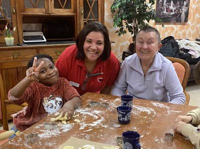 Zwei Frauen und zwei Kinder sitzen an einem Tisch und backen Plätzchen. Der Tisch ist mit Mehl bedeckt, man sieht Teig und Ausstechformen. Alle lachen in die Kamera.