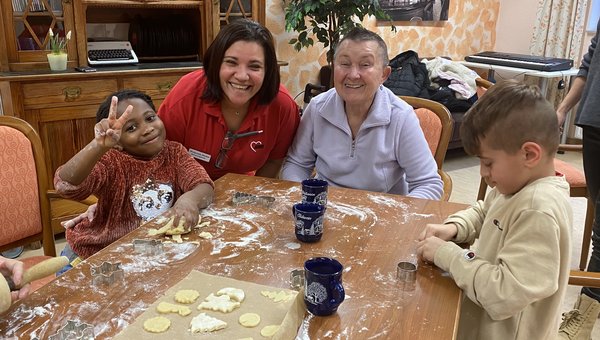 Zwei Frauen und zwei Kinder sitzen an einem Tisch und backen Plätzchen. Der Tisch ist mit Mehl bedeckt, man sieht Teig und Ausstechformen. Alle lachen in die Kamera.