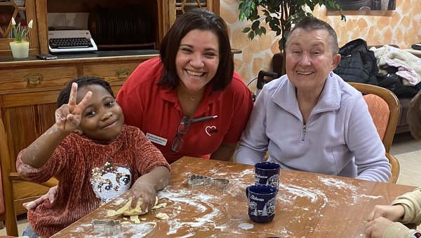 Zwei Frauen und zwei Kinder sitzen an einem Tisch und backen Plätzchen. Der Tisch ist mit Mehl bedeckt, man sieht Teig und Ausstechformen. Alle lachen in die Kamera.