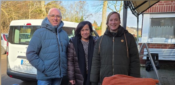 Das Bild zeigt oben zwei Frauen und einen Mann, die in die Kamera schauen. Unten ist ein Wagen mit Kartons, Tüten und Wolldecken.