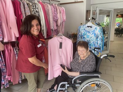 Auf dem Bild zeigt eine Frau einer Seniorin im Rollstuhl eine Jacke. Im Hintergrund stehen Kleiderständer.