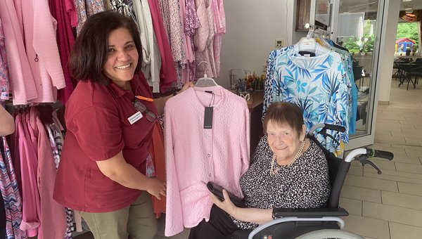 Auf dem Bild zeigt eine Frau einer Seniorin im Rollstuhl eine Jacke. Im Hintergrund stehen Kleiderständer.
