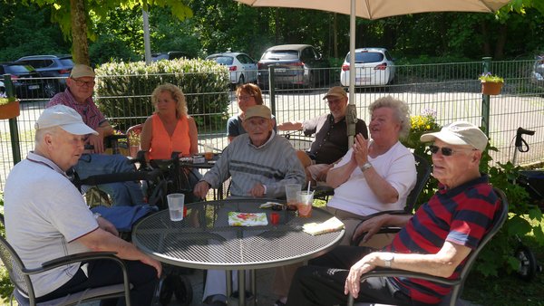Auf dem Foto ist eine Gruppe von Menschen zu sehen, die bei Sonnenschein auf einer Terrasse sitzt. Eine Seniorin klatscht zur Musik in ihre Hände