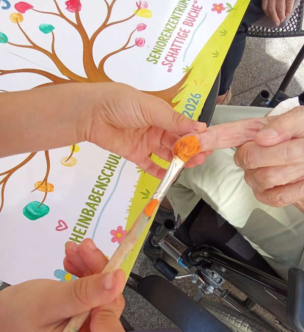 Eine Kinderhand bemalt den Finger einer älteren Person mit Farbe. Im Hintergrund zeigt ein Plakat zahlreiche bunte Fingerabdrücke.
