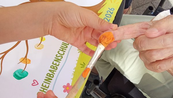 Eine Kinderhand bemalt den Finger einer älteren Person mit Farbe. Im Hintergrund zeigt ein Plakat zahlreiche bunte Fingerabdrücke.