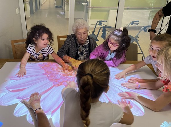 Eine Seniorin sitzt gemeinsam mit mehreren Kindern an einem Tisch. Staunend betrachten sie eine große, leuchtende Blume, die von der Tovertafel auf die Tischoberfläche projiziert wird.