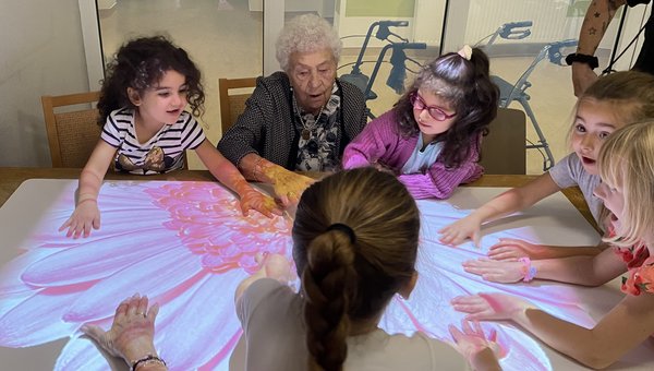 Eine Seniorin sitzt gemeinsam mit mehreren Kindern an einem Tisch. Staunend betrachten sie eine große, leuchtende Blume, die von der Tovertafel auf die Tischoberfläche projiziert wird.