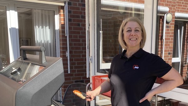 Eine Frau steht am Grill und hält in einer Hand eine Zange, mit der sie ein Grillwürstchen wendet.