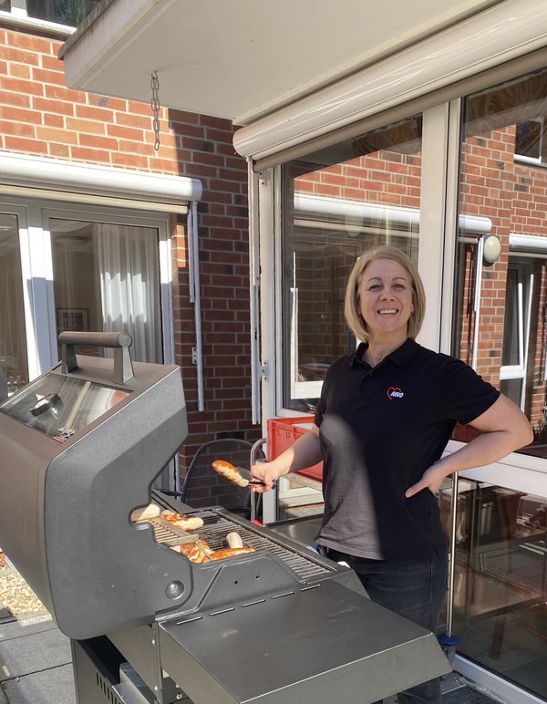 Eine Frau steht am Grill und hält in einer Hand eine Zange, mit der sie ein Grillwürstchen wendet.