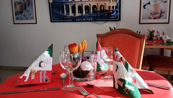 Im Vordergrund ein schön gedeckter Tisch mit Weingläsern und Servietten, die ein italienisches Muster tragen. Im Hintergrund ist ein Bild des Kolosseum in Rom zu sehen.