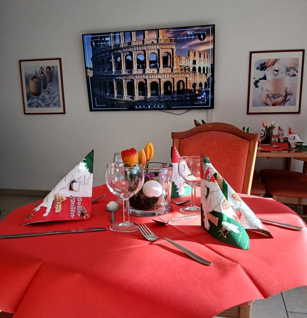 Im Vordergrund ein schön gedeckter Tisch mit Weingläsern und Servietten, die ein italienisches Muster tragen. Im Hintergrund ist ein Bild des Kolosseum in Rom zu sehen.