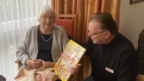 Ein Mann kniet vor einer Seniorin, die einen Adventskalender in der Hand hält und in die Kamera lächelt.