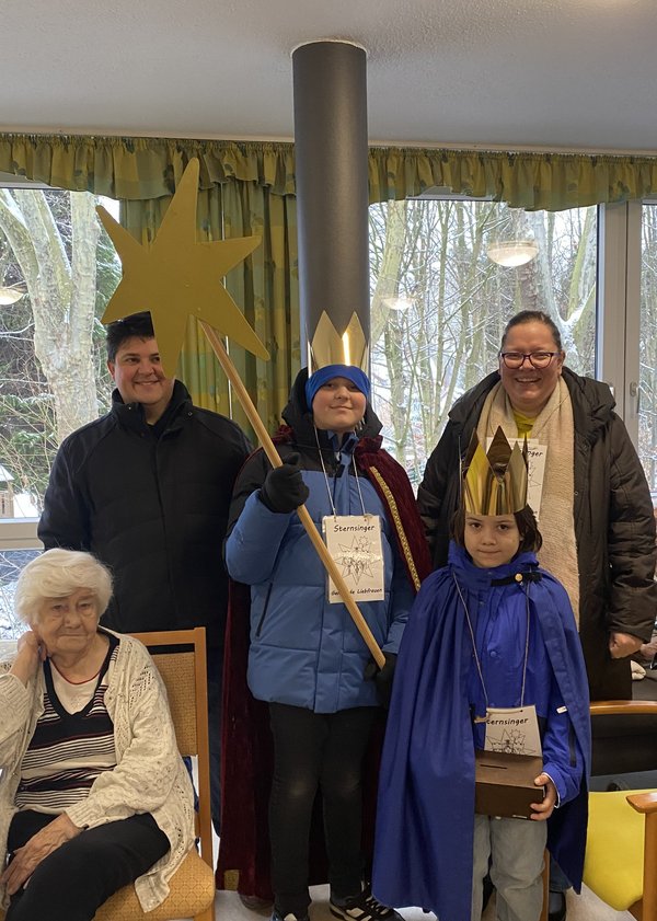Auf dem Bild ist eine kleine Gruppe von Menschen zu sehen. Zwei Kinder sind als Sternsinger verkleidet, sie tragen Umhänge, Kronen und halten einen Stern bei sich.