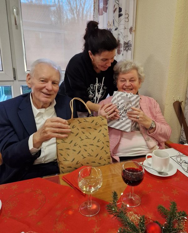 Eine Frau überreicht Geschenke an ein älteres Ehepaar, beide lachen in die Kamera.