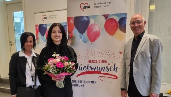 Auf dem Bild sieht man drei Personen vor einem Aufsteller mit der Aufschrift „Herzlichen Glückwunsch zur bestandenen Prüfung“ stehen. Eine der Frauen hält einen Blumenstrauß in der Hand.