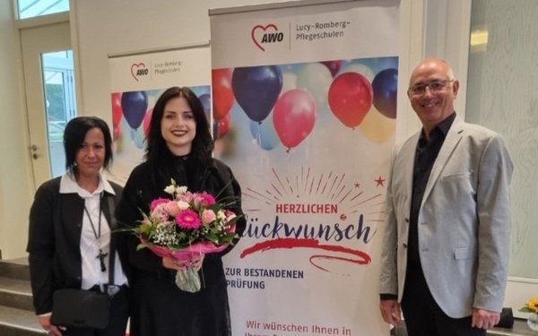 Auf dem Bild sieht man drei Personen vor einem Aufsteller mit der Aufschrift „Herzlichen Glückwunsch zur bestandenen Prüfung“ stehen. Eine der Frauen hält einen Blumenstrauß in der Hand.