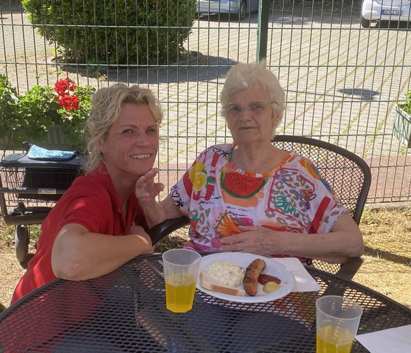 Zwei Frauen sitzen auf einer Terrasse und schauen lächelnd in die Kamera. Vor der Seniorin steht ein Teller mit Grillwurst und Kartoffelsalat.