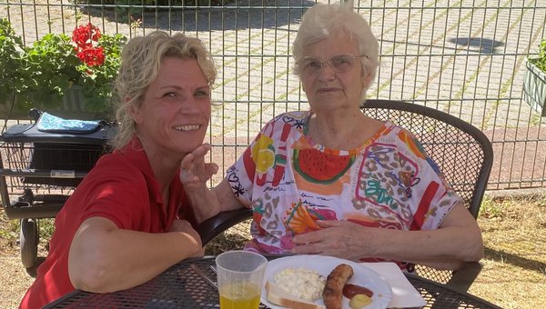 Zwei Frauen sitzen auf einer Terrasse und schauen lächelnd in die Kamera. Vor der Seniorin steht ein Teller mit Grillwurst und Kartoffelsalat.