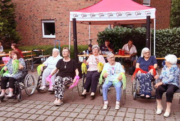 Eine Gruppe Seniorinnen sitzt nebeneinander und führt gemeinsam einen Sitztanz auf. An ihren Handgelenken tragen sie bunte Tücher.