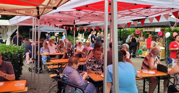 Sommerfest im Freien mit Bierzeltgarnituren und Pavillons. Menschen sitzen zusammen, essen und trinken. Bunte Luftballons und Wimpel schmücken das Festgelände.