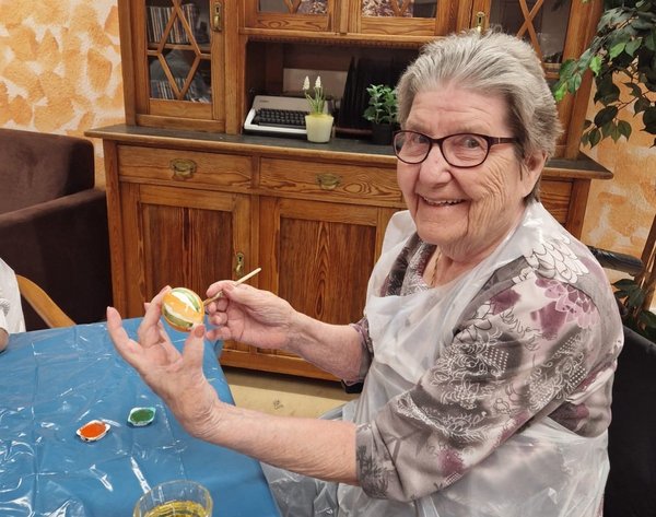 Eine Seniorin bemalt ein Osterei und lächelt dabei in die Kamera.